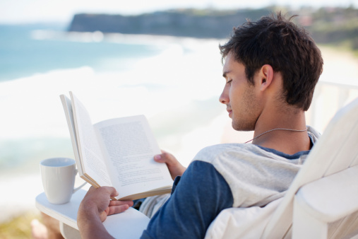 Man with coffee cup reading book in deck chair overlooking ocean